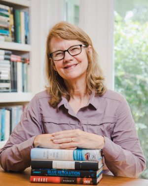 Cynthia Barnett at desk with her hands on a stack of books and shelves with books in the background