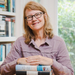 Cynthia Barnett at desk with her hands on a stack of books and shelves with books in the background