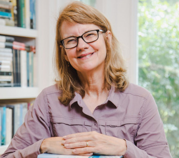 Cynthia Barnett at desk with her hands on a stack of books and shelves with books in the background