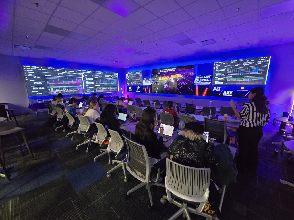 A group of students sits at a row of tables in the foreground with laptops open in front of them. Another student stands just off to the right, wearing a black and white referee jersey. The background features a row of blueish video wall screens displaying charts, graphs, and a football game.