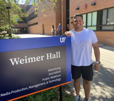 Luis Rodrigues stands next to the blue and black sign for Weimer Hall