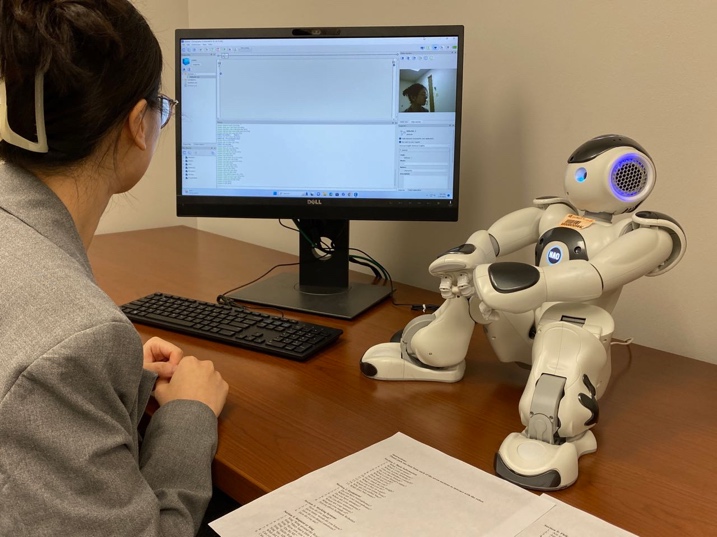 A student seated at a desktop computer with a small humanoid robot sitting on the desk. 