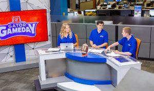 Three student news anchors sitting behind a glass and steel newsroom desk.